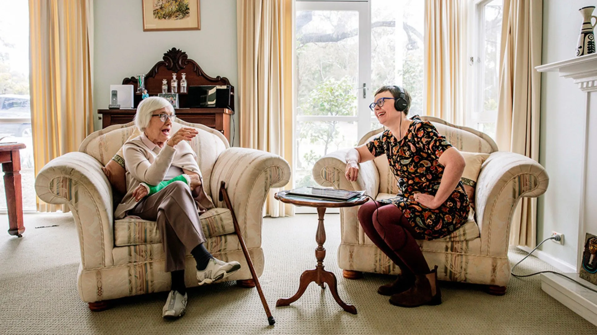 An elderly woman sits in an armchair talking to a younger woman wearing headphones in a cozy, sunlit living room.