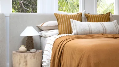 Cozy bedroom with beige and mustard pillows, throw blanket, and side table with lamp and decorative items.