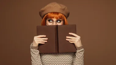 Red-haired woman with glasses wearing a beret, holding a book upright, peeking over it against a brown background.