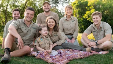 A family in khaki shirts from Australia Zoo sits on a picnic blanket on grass with trees in the background.
