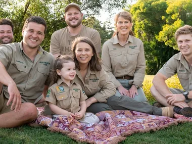 A family in khaki shirts from Australia Zoo sits on a picnic blanket on grass with trees in the background.