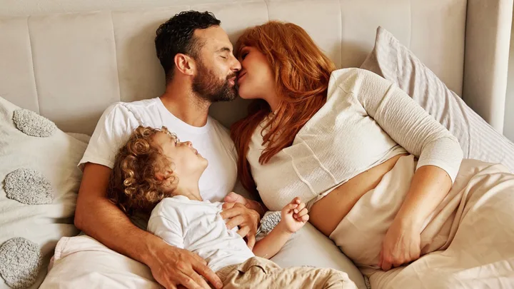 Family cuddling in bed; parents kiss while child smiles, lying between them.