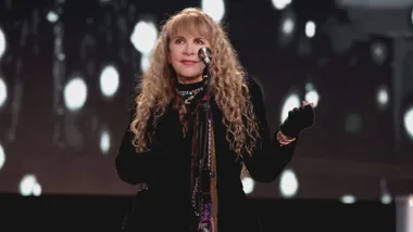 Singer with curly blonde hair performs on stage, holding a microphone, wearing a black outfit with necklaces.