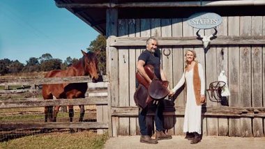 Man and woman stand with a horse and a parrot outside "The Stables" wooden barn in a rural setting.