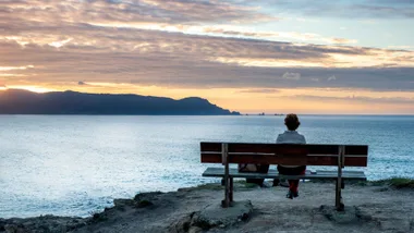 Person sitting on a bench overlooking the ocean at sunset, with mountains in the distance under a cloudy sky.