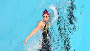 Swimmer in a cap and goggles performs a backstroke in a pool, creating splashes in the clear blue water.