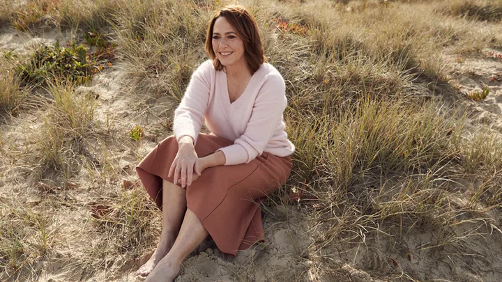 A woman sits smiling on sand dunes, wearing a pink sweater and skirt, surrounded by tall grass.