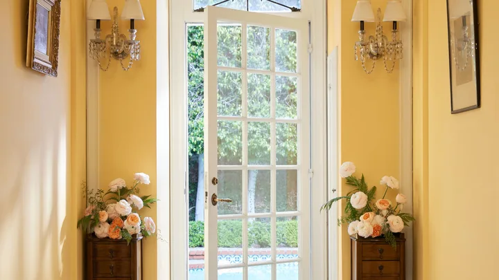 Open white French doors leading to a garden, with vases of peach and white flowers on either side in a yellow hallway.