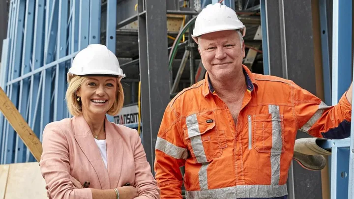 Two people wearing hard hats, smiling at a construction site; one in a pink blazer, the other in an orange safety shirt.