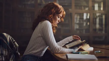 Woman with red hair reading a book at a library table surrounded by papers and pens.