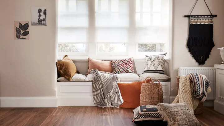 Cozy window nook with cushioned seating, pillows, blankets, and wall decor in neutral tones.