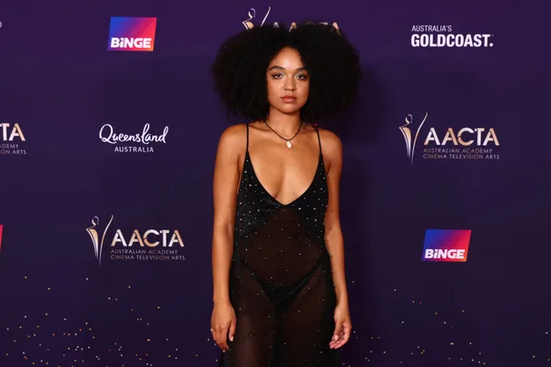 A person in a black dress stands in front of a backdrop with AACTA and Binge logos at an event.