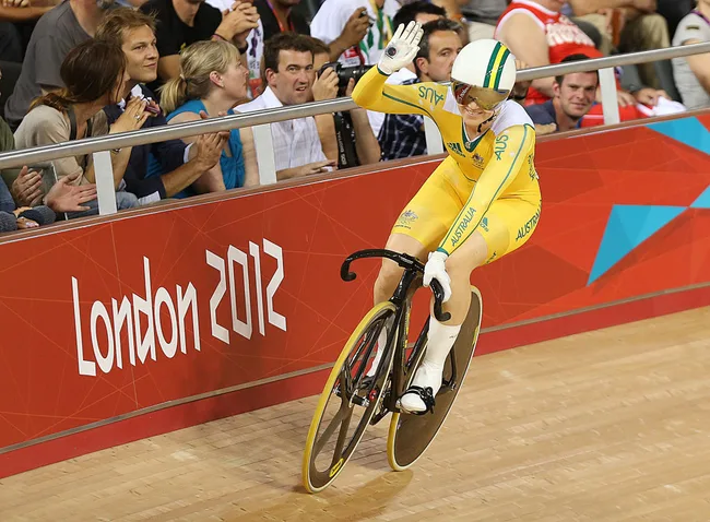 Cyclist in yellow Australian uniform waves during London 2012 Olympics track event with cheering audience.