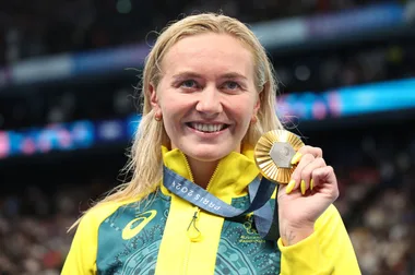 Australian athlete smiling and holding a gold medal, wearing a green and yellow jacket, with a blurred crowd in background.