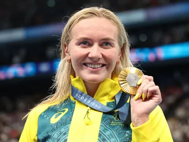 Australian athlete smiling and holding a gold medal, wearing a green and yellow jacket, with a blurred crowd in background.