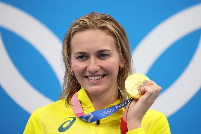 Athlete smiles while holding a gold medal at the Tokyo 2020 Olympics, wearing a yellow outfit.