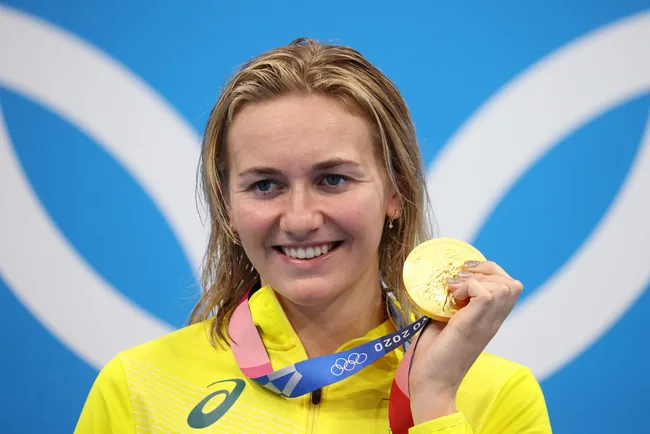 Athlete smiles while holding a gold medal at the Tokyo 2020 Olympics, wearing a yellow outfit.