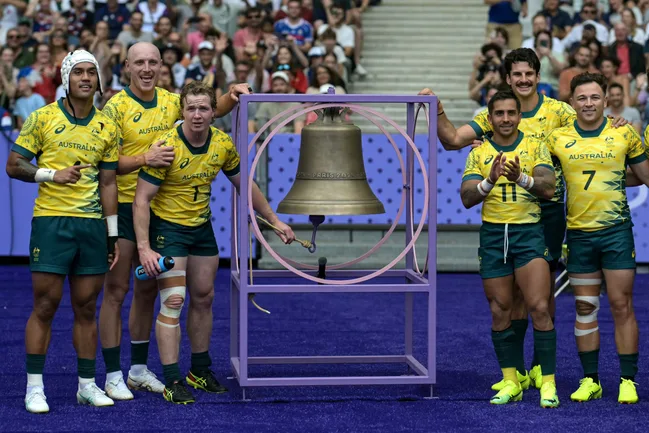 Australian rugby players stand around a bell on a field, wearing yellow and green uniforms, with a cheering crowd behind.