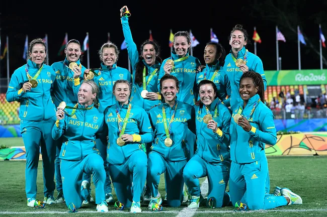 Australian women's rugby sevens team celebrating with gold medals at Rio 2016 Olympics, all in teal uniforms.