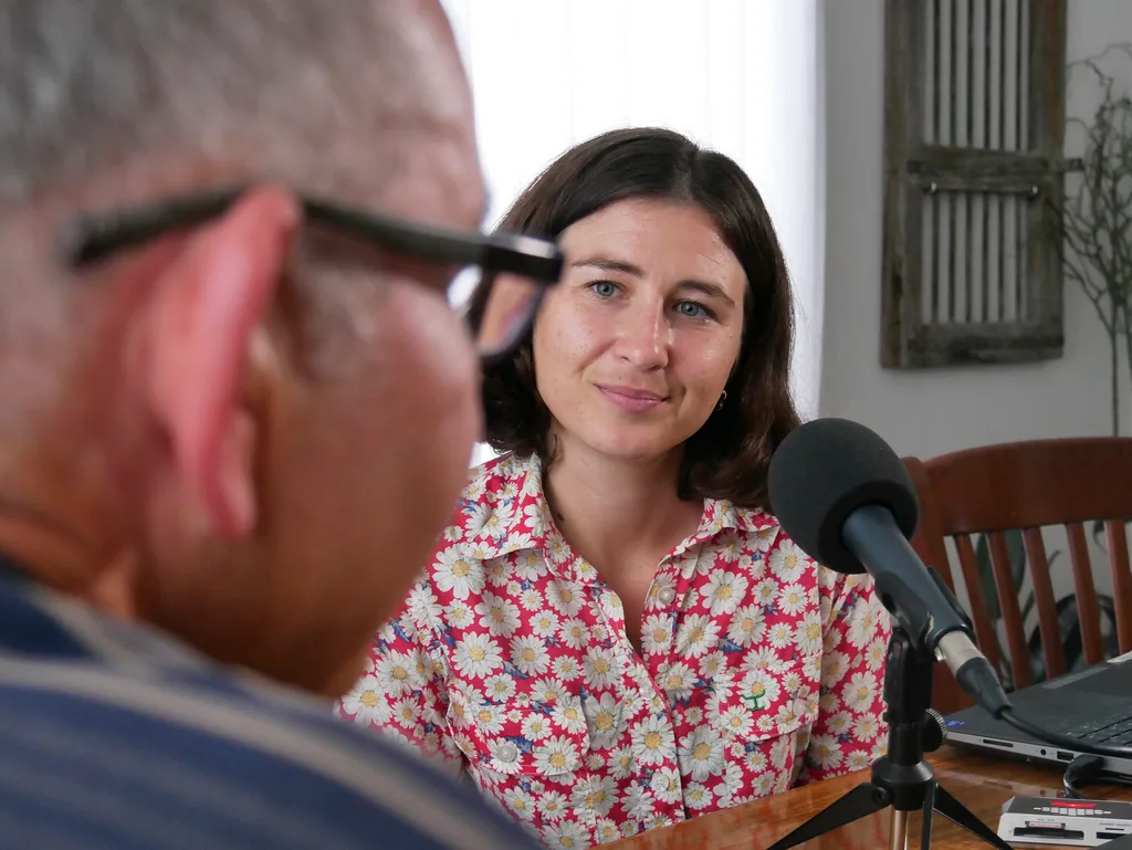 A woman sits at a table with a microphone, interviewing a man.