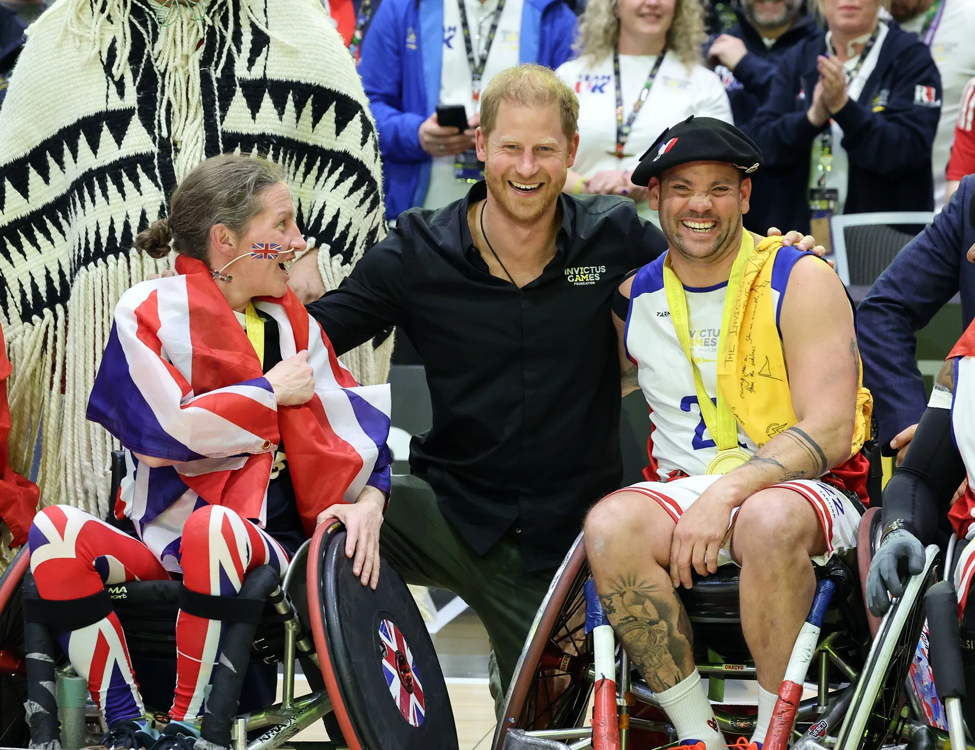 Prince Harry at the Invictus Games wearing a black polo shirt kneels between two people in wheelchairs, one draped in the UK flag, the other wearing a beret.
