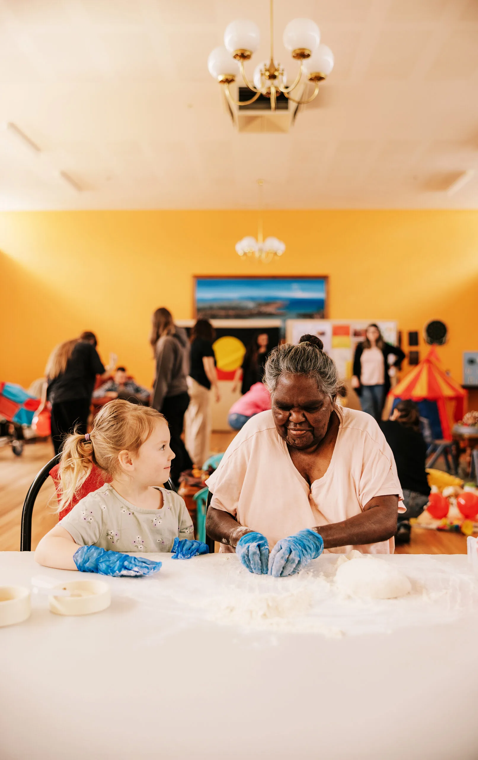 A young girl and an older Aboriginal woman sit together at a table making damper as part of an intergenerational playgroup.