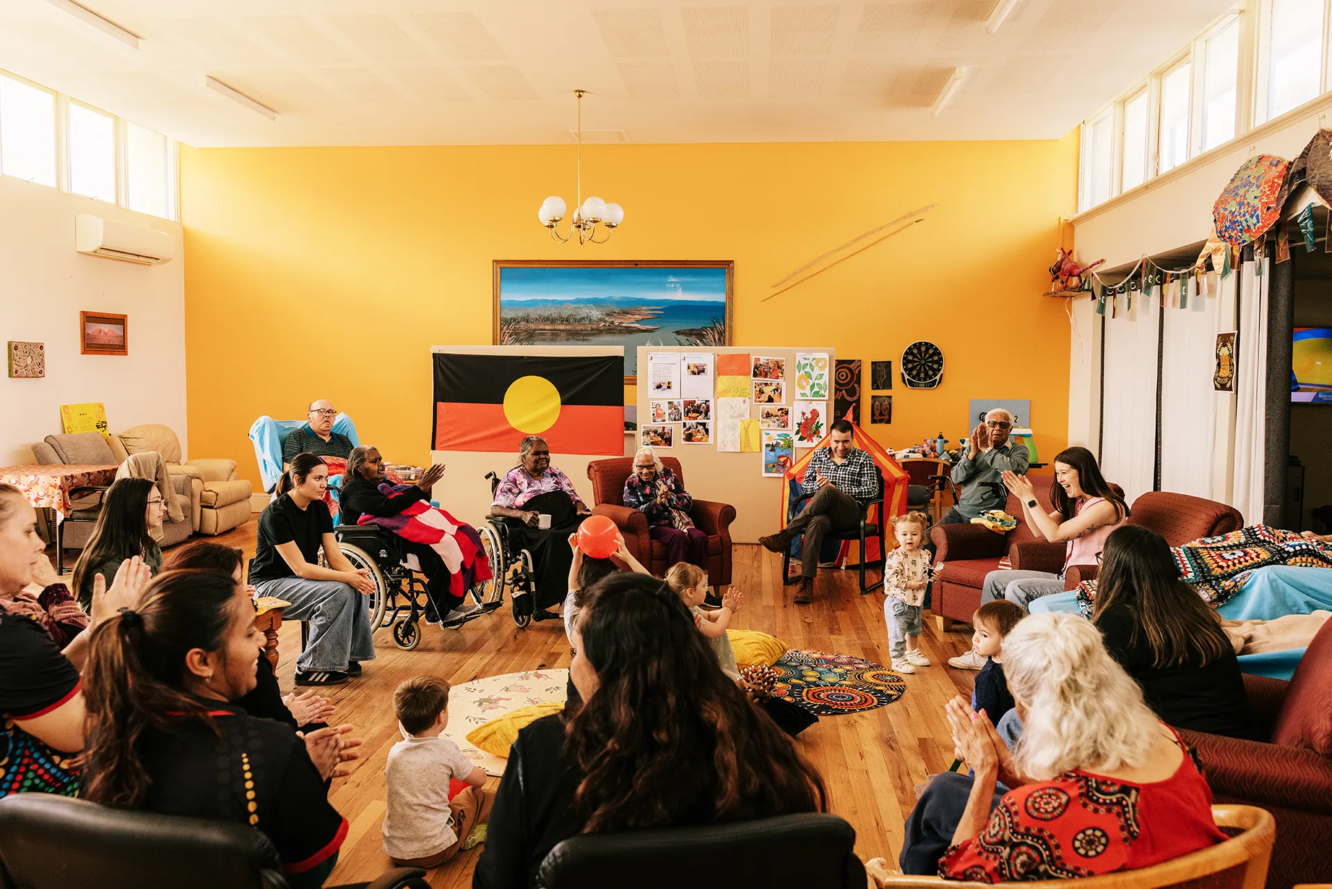 A group of people sat in a circle, clapping hands and singing at an intergenerational playgroup.