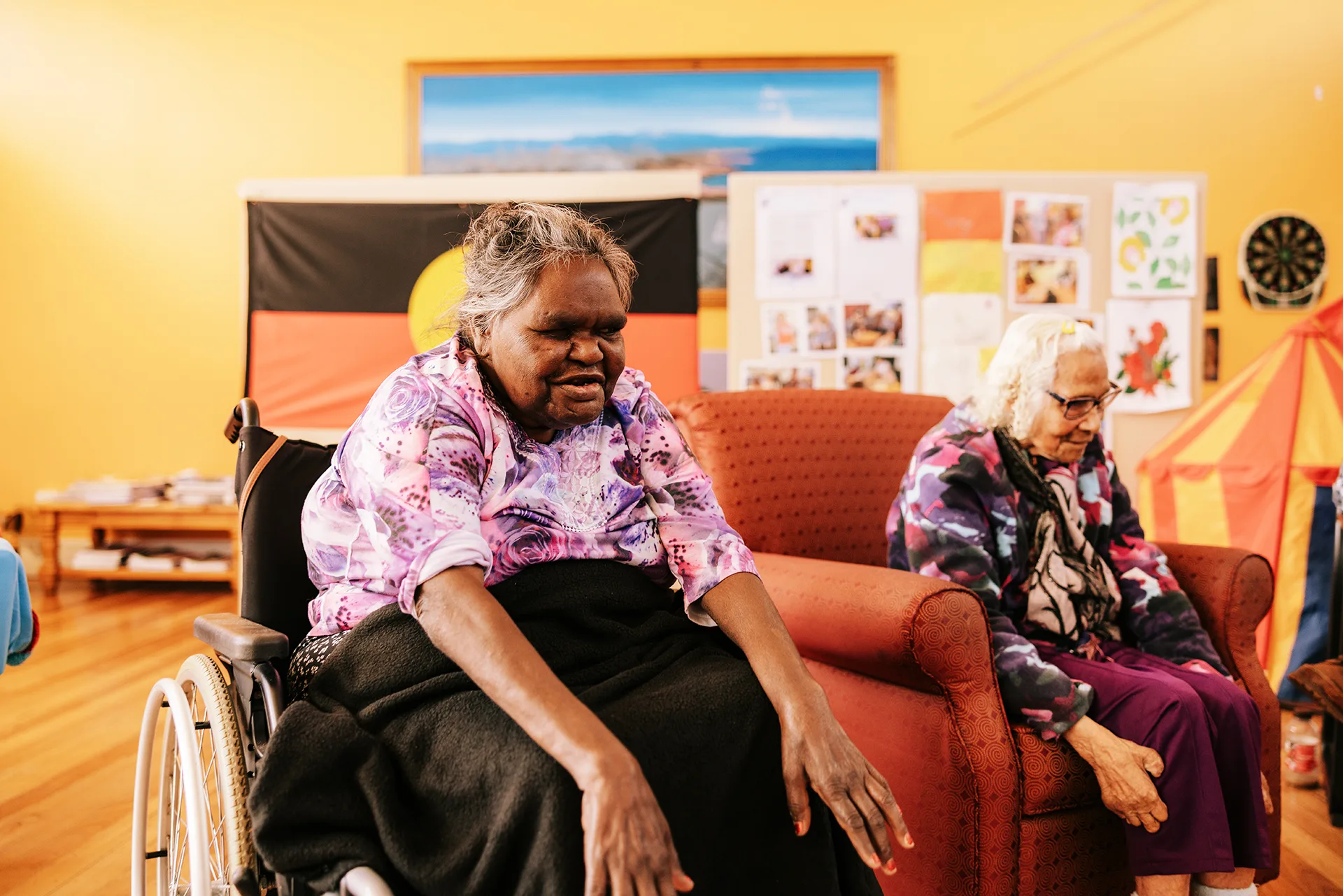 Two elderly women sitting in a recreation centre with an Aboriginal flag behind them.