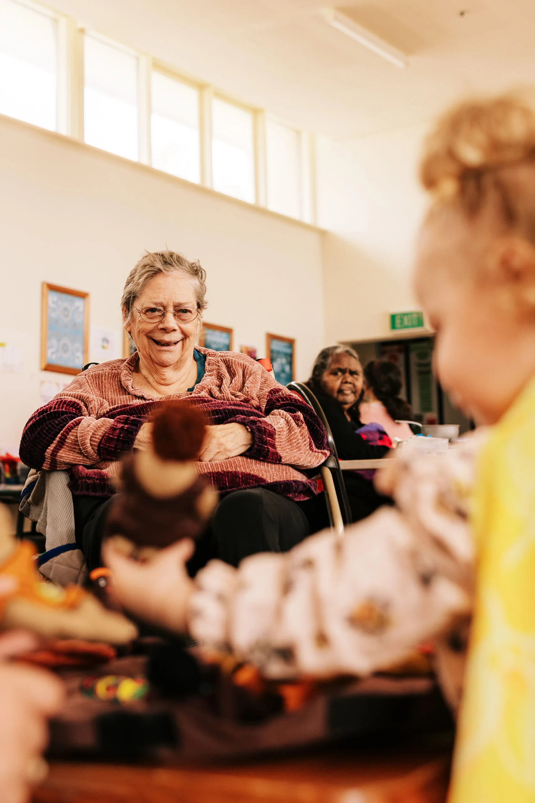 An older woman sits in a chair, smiling at a girl playing with dolls.