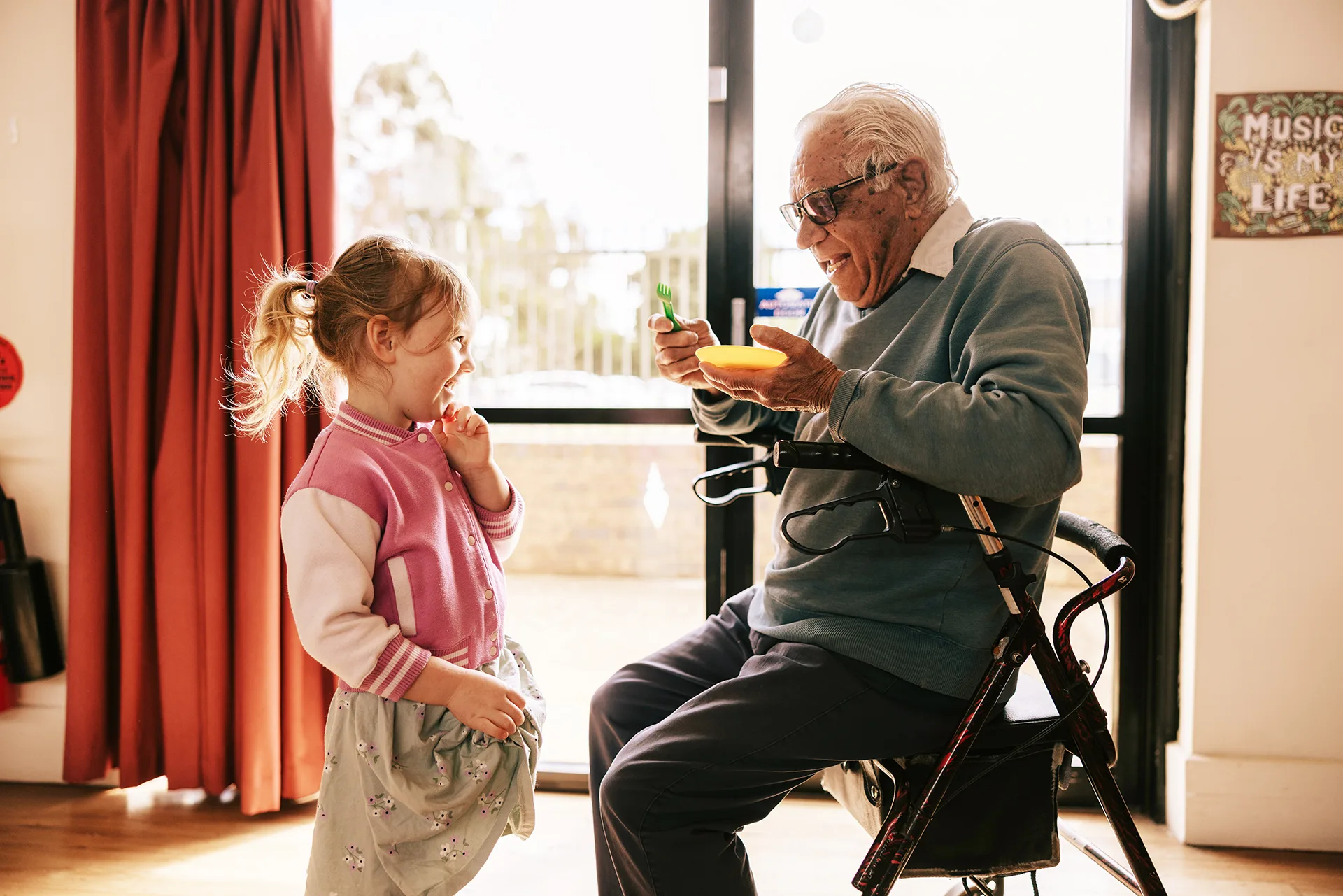 An older man sitting on a walker and a young girl laughing together at an intergenerational playgroup.