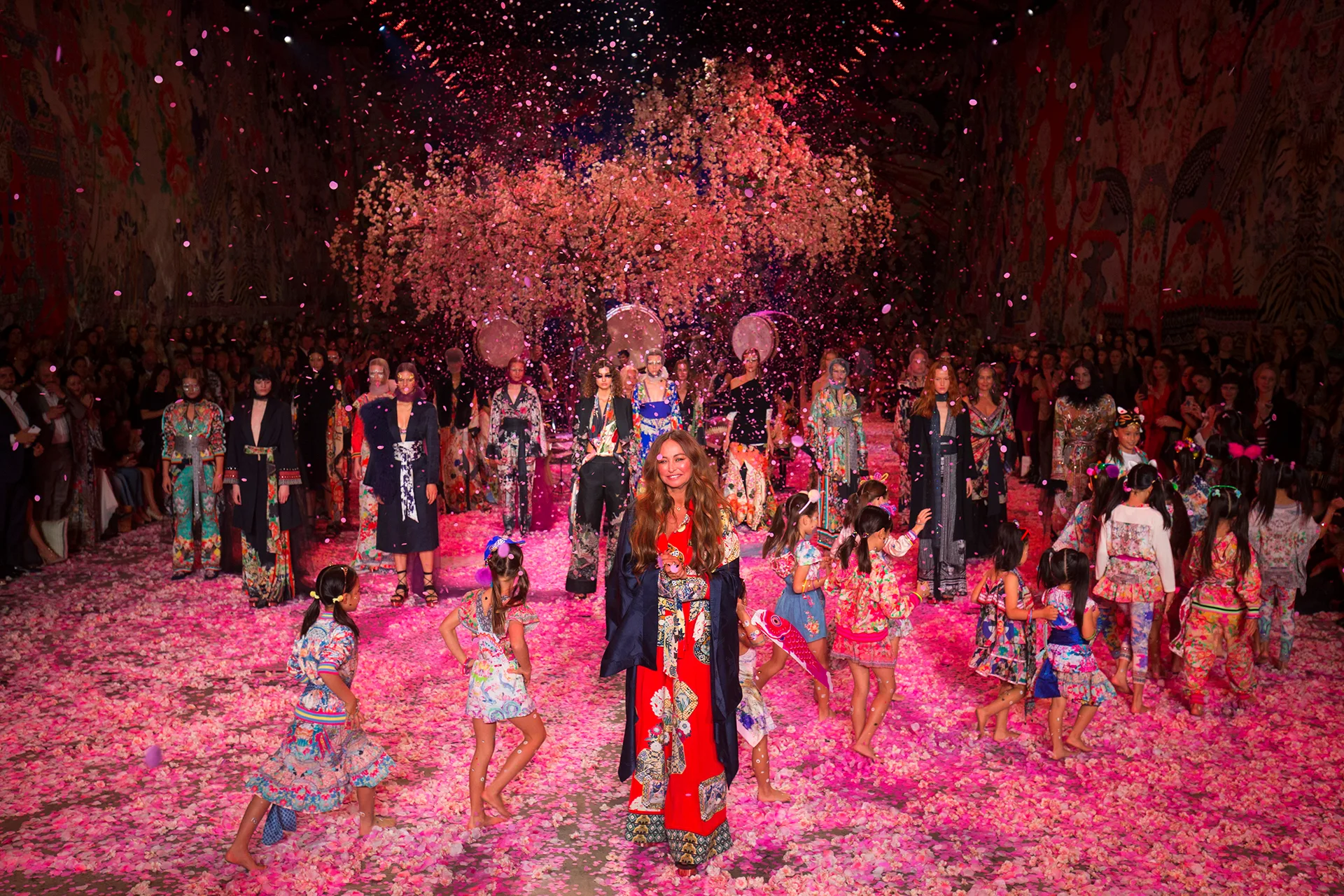 Several adults and children standing on a floor scattered with colourful flowers