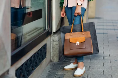 Woman holding a large handbag, wearing jeans and white espadrilles, standing by a shop window on a city street.