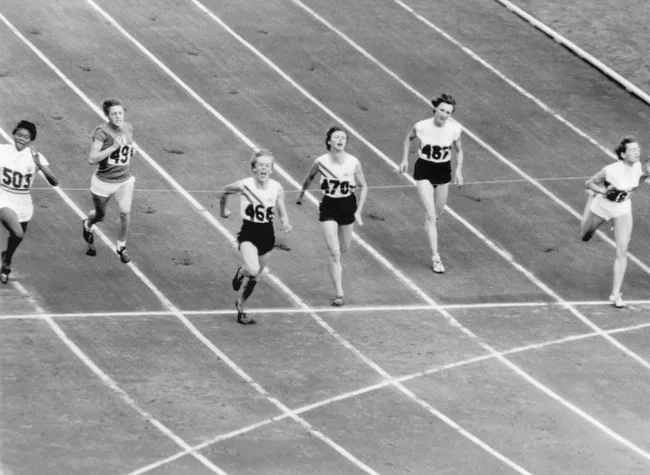 Black and white photo of female athletes running on a track, nearing the finish line in a race.