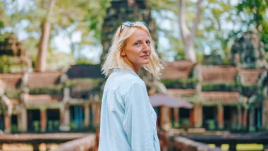 A woman with blonde hair wearing a light shirt stands in front of ancient temple ruins, looking back at the camera.