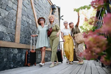 Four women dressed stylishly, smiling and walking with luggage on a wooden path next to a stone building.