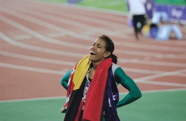 A smiling athlete draped in multiple flags stands on a track, celebrating.