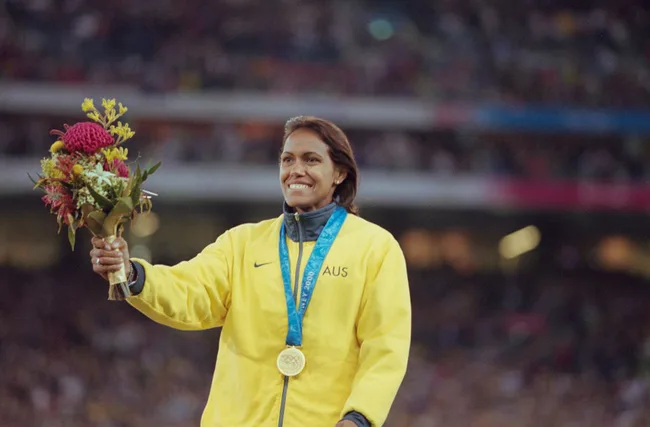 Australian athlete with gold medal and flowers, wearing yellow jacket, smiling on podium at Sydney 2000 Olympics.