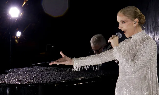 A woman in a sparkling dress sings into a microphone on stage, with a spotlight behind her and a pianist in the background.