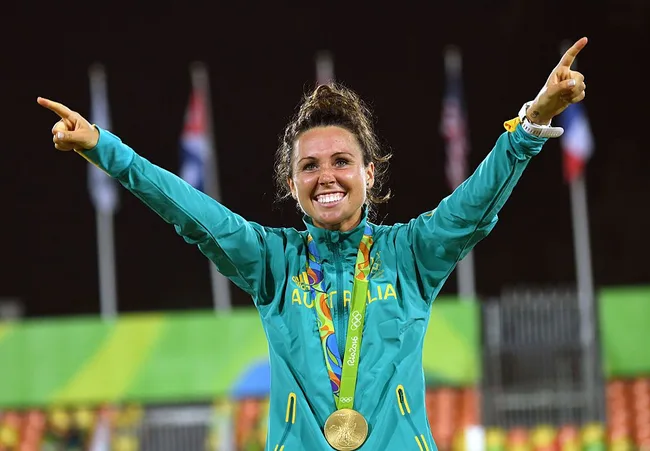 Australian athlete in a green jacket celebrates with a gold medal at an event, arms raised, flags in the background.