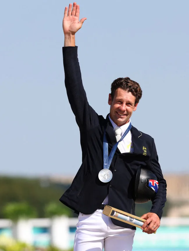 Athlete in a navy jacket and white pants waves, holding a helmet and medal, against a clear blue sky background.