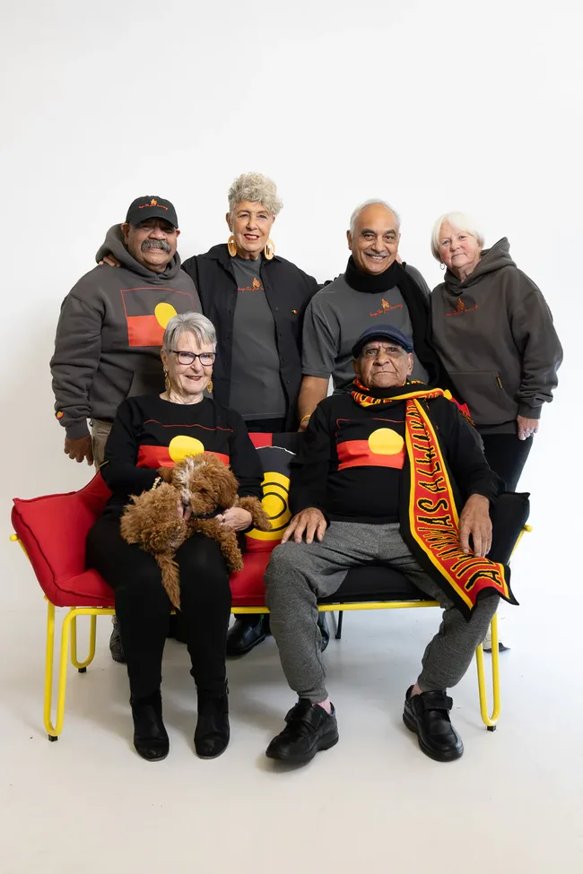 Group of six people, some seated, wearing Aboriginal flag clothing, with a dog, smiling against a white background.