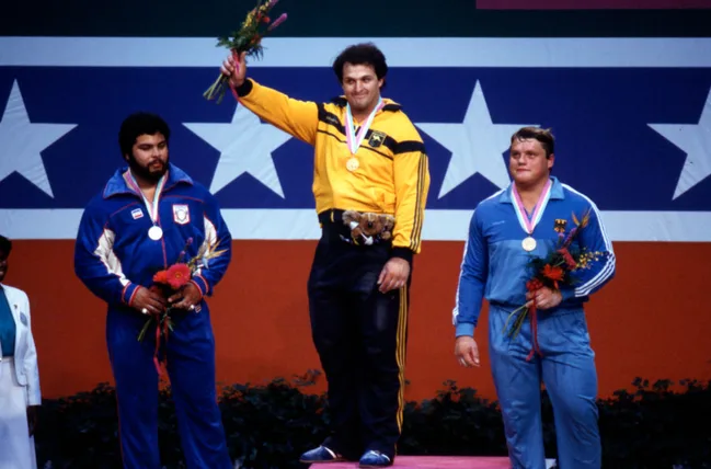Three athletes on an Olympic podium with medals and flowers, in front of a star-spangled backdrop.