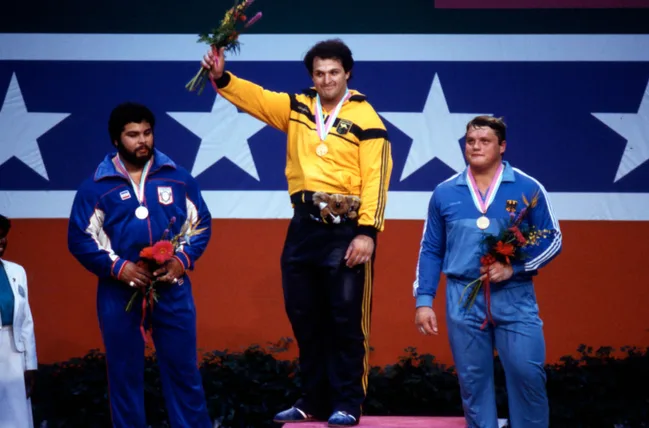 Three athletes on an Olympic podium with medals and flowers, in front of a star-spangled backdrop.