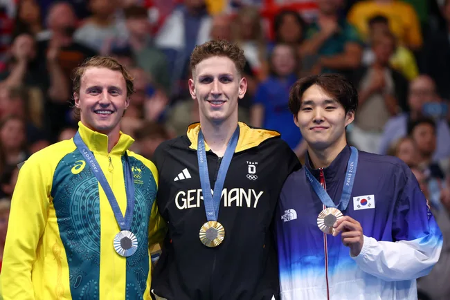 Three athletes stand on a podium, wearing medals. They represent Australia, Germany, and South Korea.