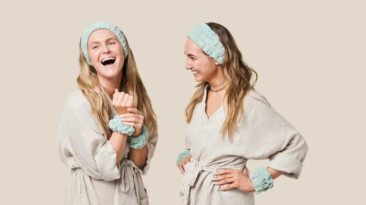 Two women wearing robes and teal headbands laughing together against a beige background.