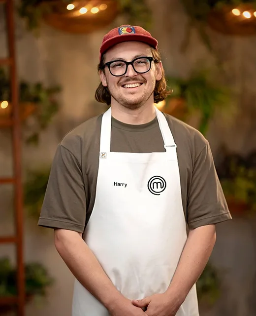 A smiling man wearing glasses, a red cap, and a "Harry" MasterChef apron stands in a warmly lit room with plants.
