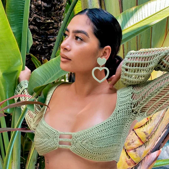 Person in a green crochet top with large heart earrings poses among tropical leaves.