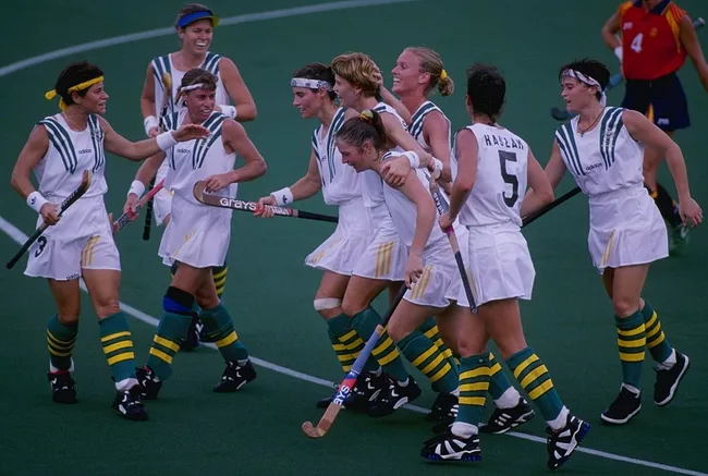 A women's field hockey team in white and green uniforms celebrates energetically on a grass field.