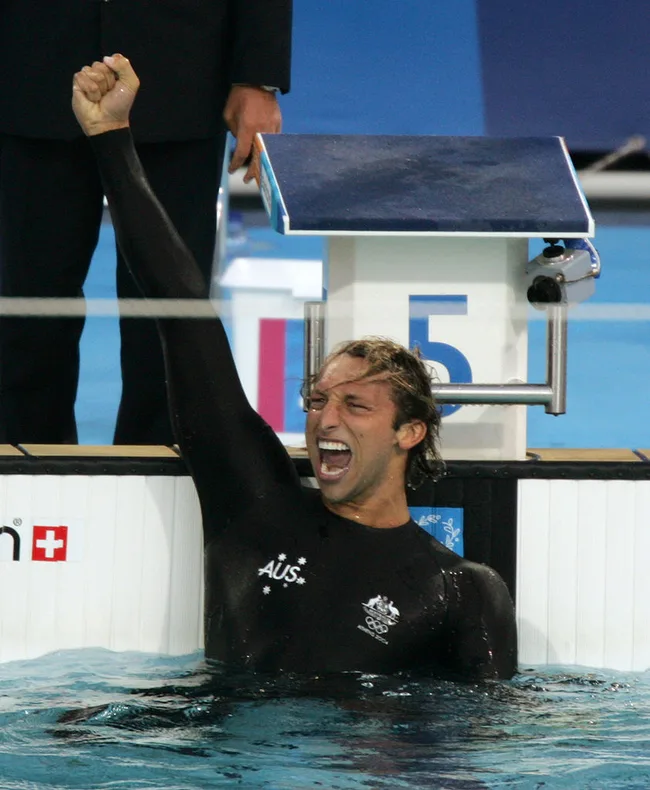 A swimmer in a black suit celebrates with a raised fist at a competition poolside.