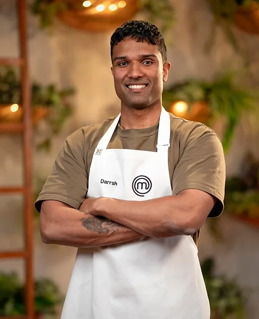 Man in a MasterChef apron smiling, standing with arms crossed in a warmly lit kitchen setting.